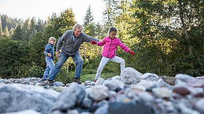 Großvater hilft zwei Kindern beim Balancieren über Steine in einem Bach, umgeben von Wald und Natur.