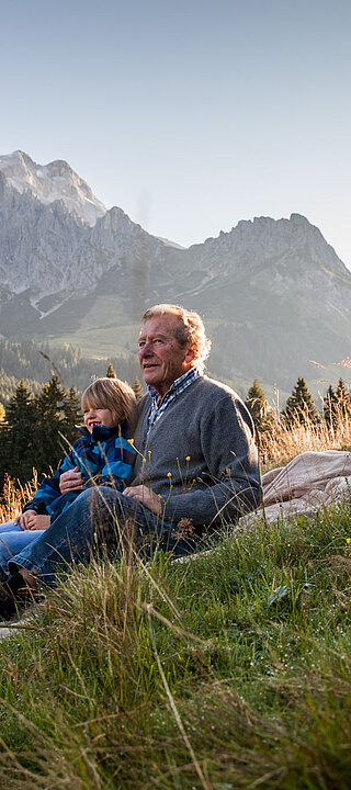 Großvater sitzt mit einem Kind auf einer Wiese und blickt in die Berglandschaft des Hochkönigs bei warmem Abendlicht.