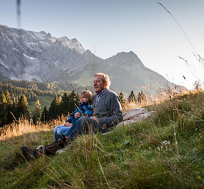 Großvater sitzt mit einem Kind auf einer Wiese und blickt in die Berglandschaft des Hochkönigs bei warmem Abendlicht.