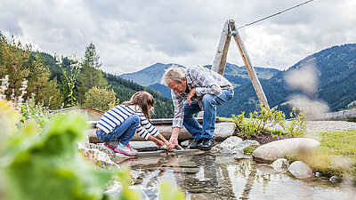 Großvater und Kind knien an einem kleinen Wasserlauf und spielen gemeinsam mit Wasser und Steinen inmitten der Natur.