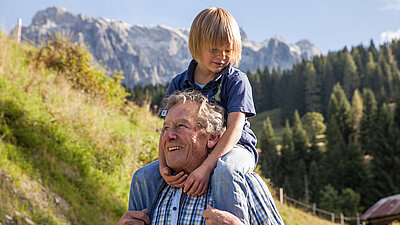 Kind sitzt auf den Schultern des Großvaters und blickt gemeinsam mit ihm in die umliegende Berglandschaft.