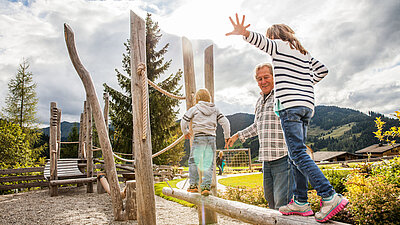 Großvater balanciert mit zwei Kindern auf einem Holzparcours am Spielplatz, im Hintergrund Berge und bewölkter Himmel.
